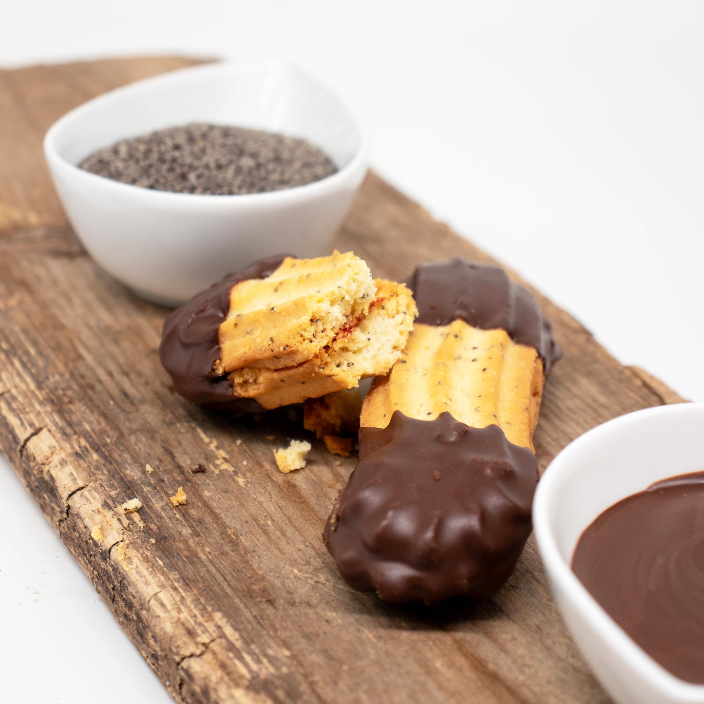 Gezeigt werden zwei länglich, hell gebackene Graumohnstangerl, deren Enden in dunkle Schokolade getaucht sind. Einer der Kekse ist in der Mitte gebrochen, sodass die butterweiche Konsistenz sichtbar wird.