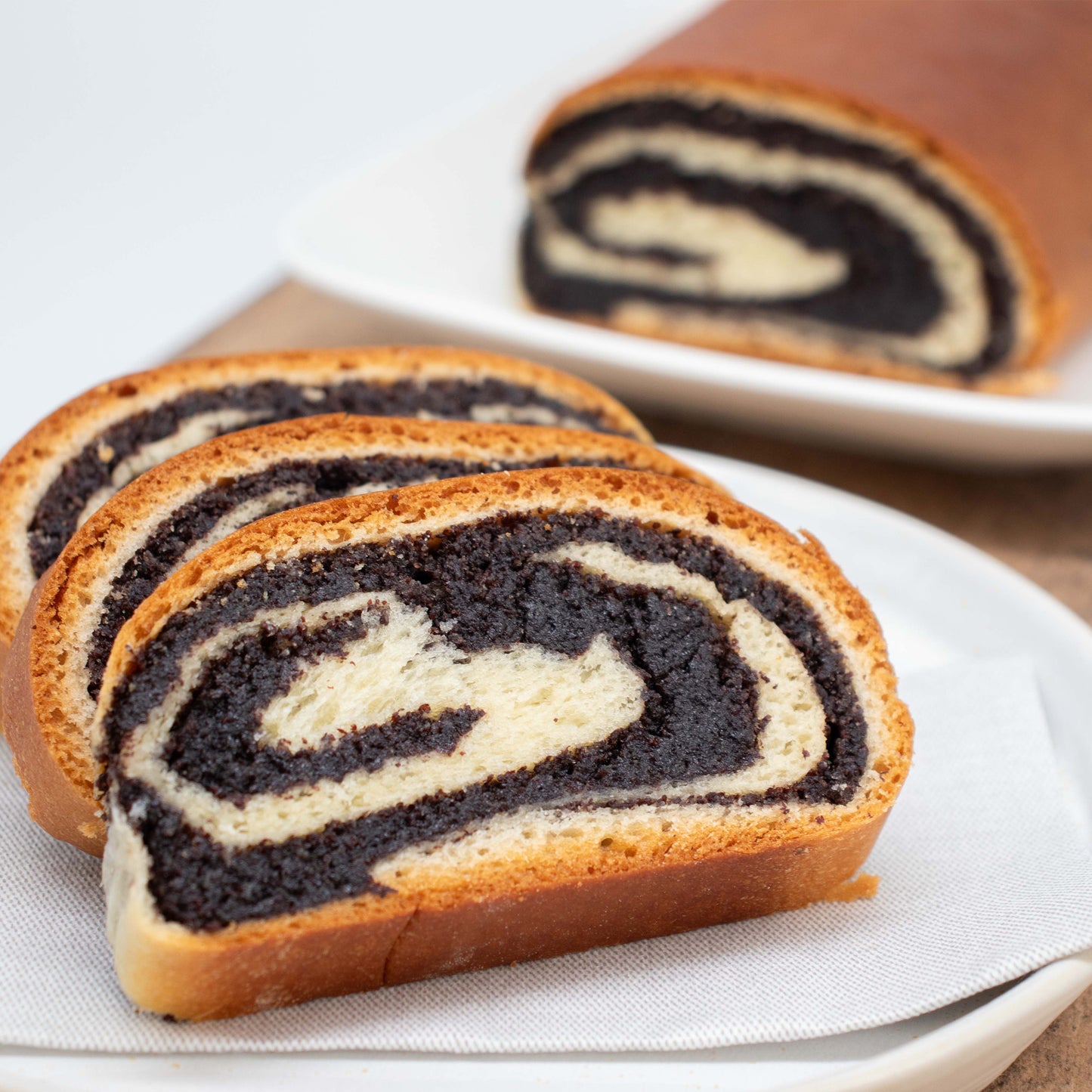 3 Scheiben Monstrudel liegen aufgeschnitten auf einem Teller. Die saftige Mohnfülle sticht ins Auge. Im Hintergrund liegt leicht verschwommen der restliche Mohnstrudel im Ganzen.