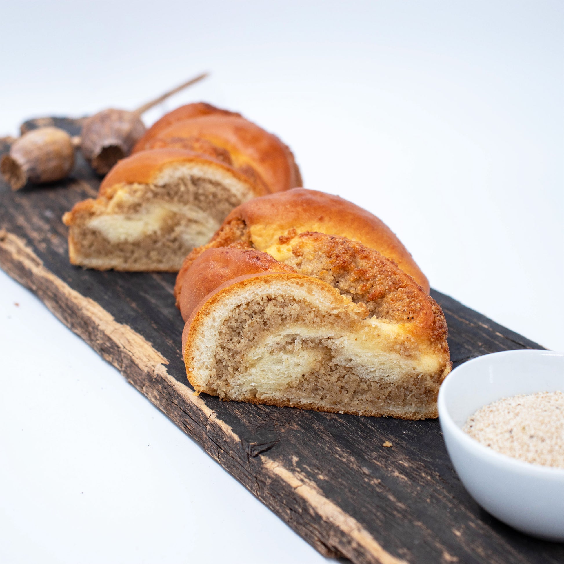 Zu sehen ist ein auseinandergeschnittener Weißmohnstriezel. Die zwei Hälften liegen nebeneinander und man sieht deutlich die saftige Weißmohnfülle. Im Hintergrund leicht verschwommen sieht man Weißmohnkapseln und im Vordergrund eine weiße Schale mit Weißmohnsamen. 