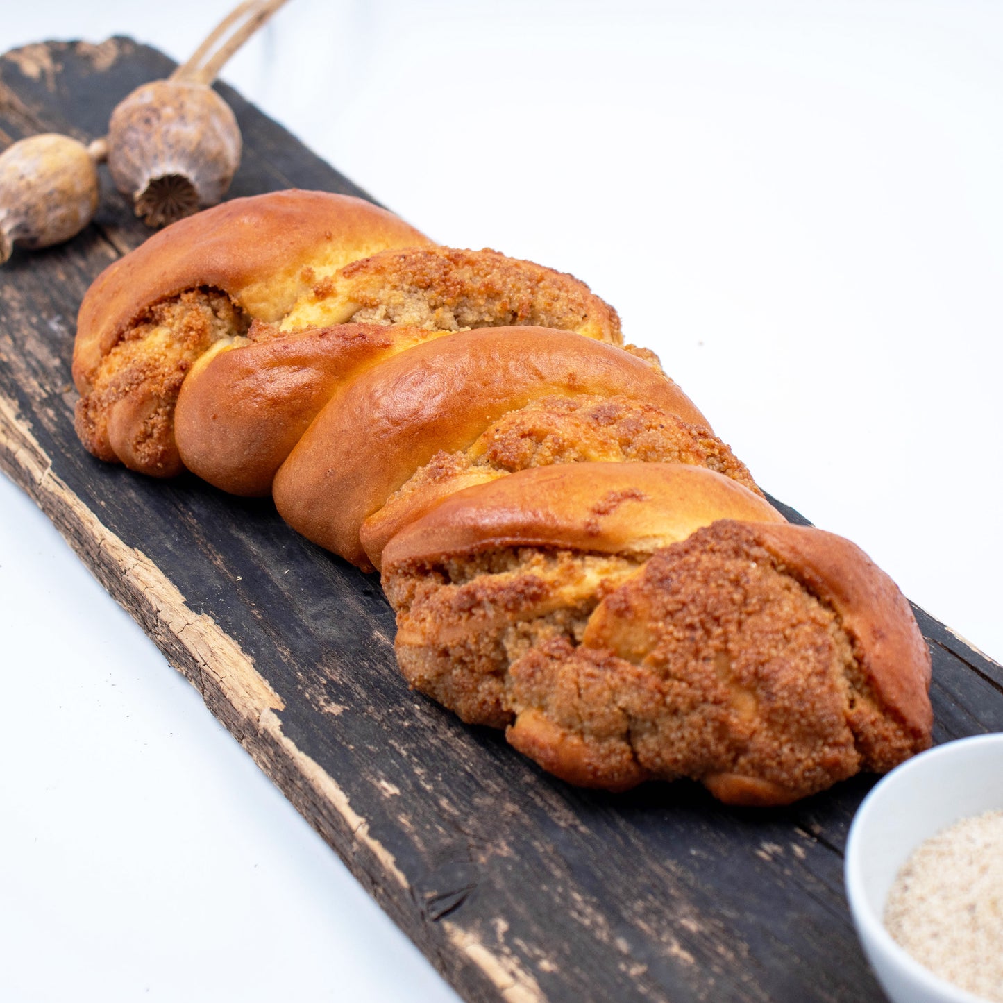 Zu sehen ist ein Weißmohnstriezel. Im Hintergrund leicht verschwommen sieht man Weißmohnkapseln und im Vordergrund eine weiße Schale mit Weißmohnsamen. 