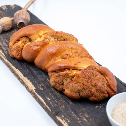 Zu sehen ist ein Weißmohnstriezel. Im Hintergrund leicht verschwommen sieht man Weißmohnkapseln und im Vordergrund eine weiße Schale mit Weißmohnsamen. 