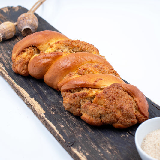 Zu sehen ist ein Weißmohnstriezel. Im Hintergrund leicht verschwommen sieht man Weißmohnkapseln und im Vordergrund eine weiße Schale mit Weißmohnsamen. 